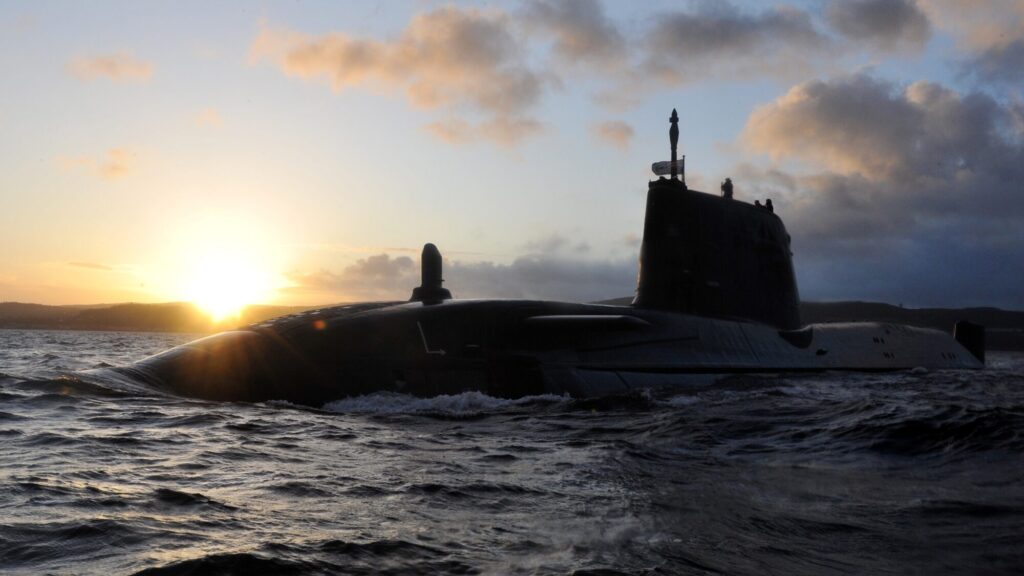 A submarine is partially submerged in the ocean at sunset, with waves around it and the sun low on the horizon.